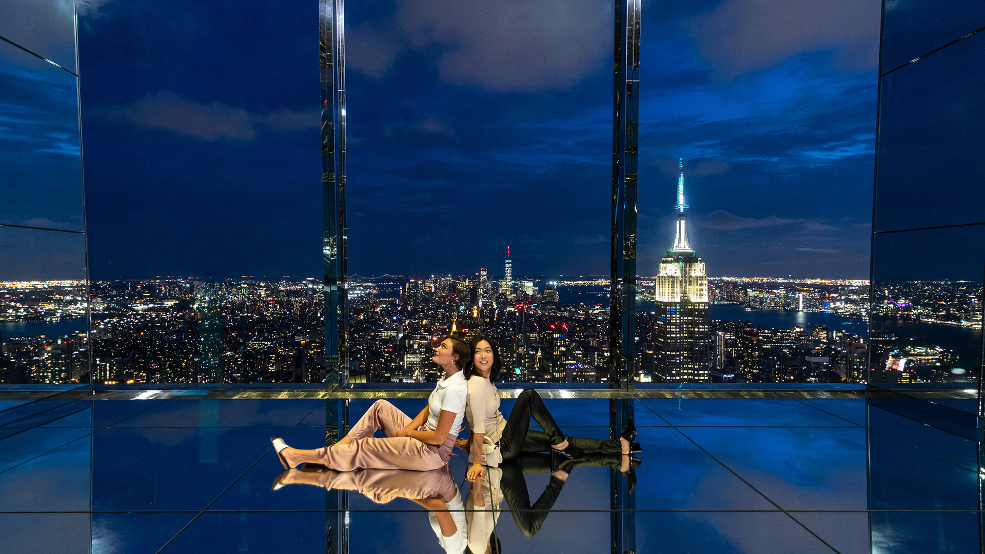 Two women, positioned back to back, absorbing the view of New York at night.