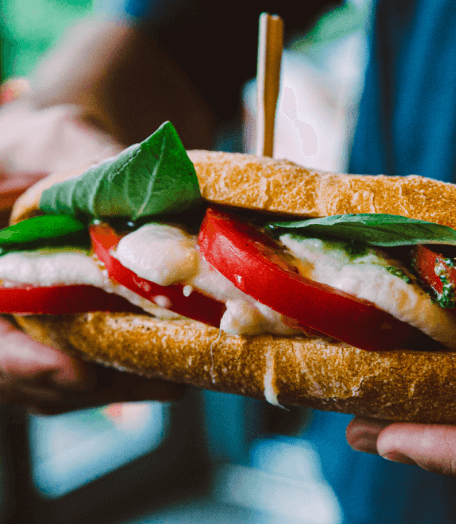 Close-up of a New York sandwich with fresh mozzarella, tomatoes, and basil, held in hand with a wooden skewer through it.